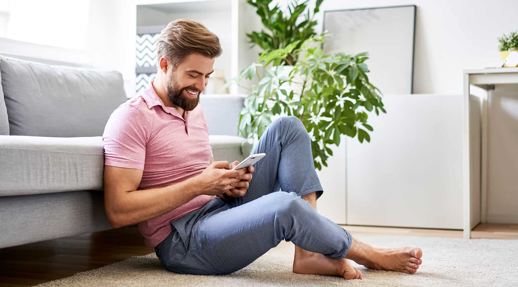 Nicely dressed man sitting on the hardwood floors of an apartment, smiling and looking at his cell phone.