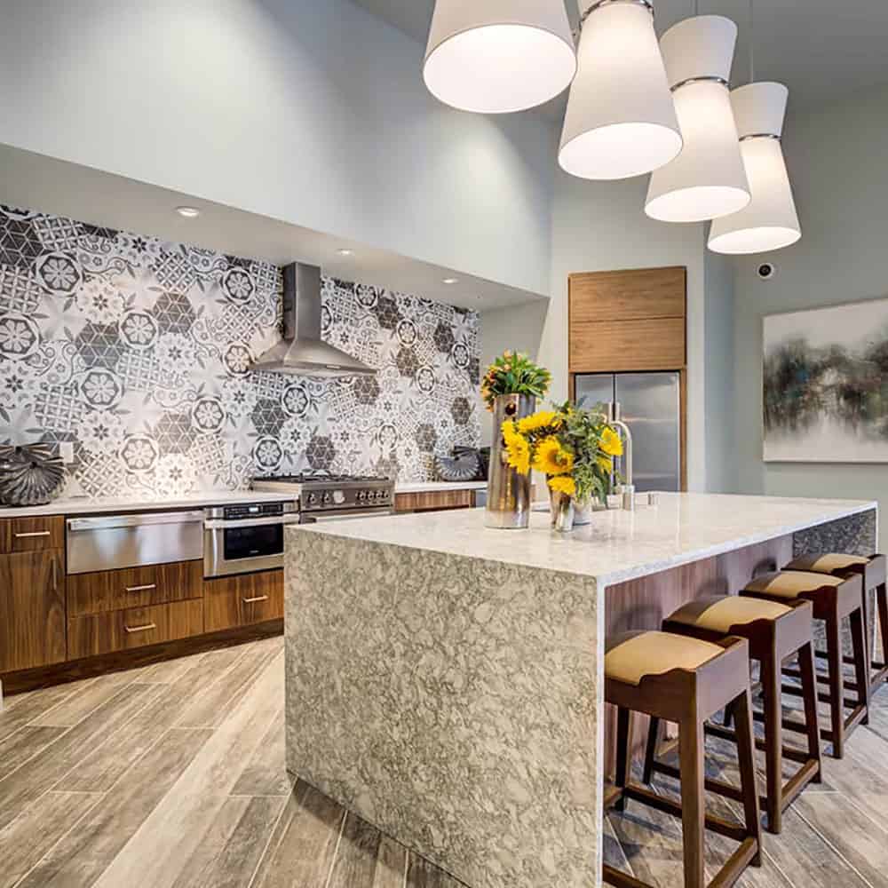 Common kitchen area in a luxury apartments' building main floor; featuring upscale steel appliances, a marble-made countertop next to high chairs.